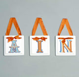 Three white square hanging ornaments with orange ribbon loops, a large decorative letter